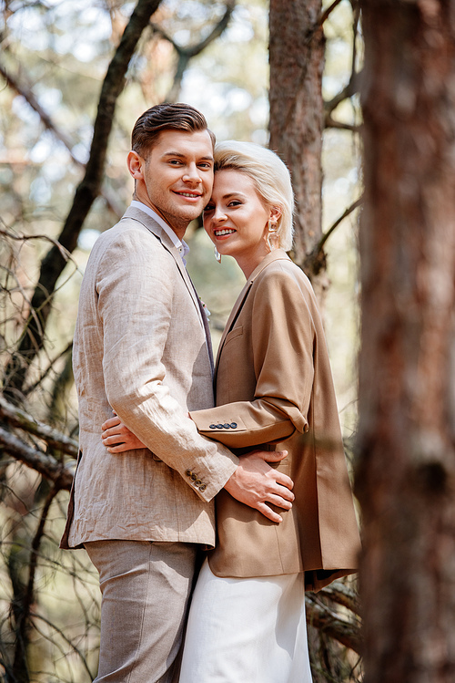stylish smiling couple in elegant jackets embracing in forest