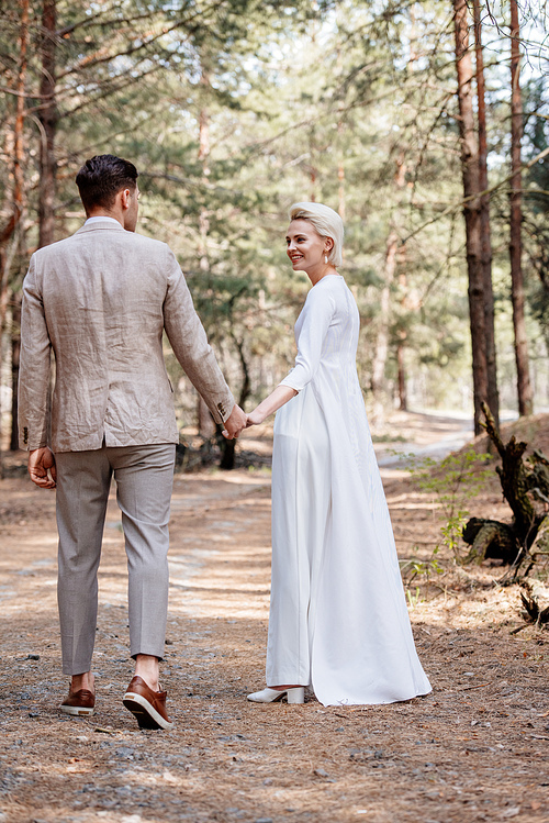 full length view of smiling just married couple holding hands and walking in forest