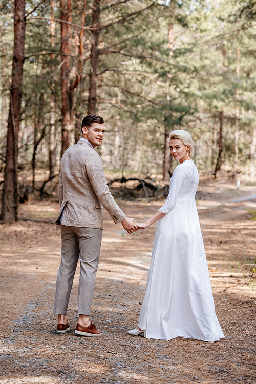 full length view of smiling just married couple holding hands in forest