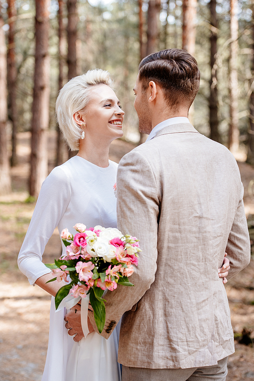 just married couple in formal wear embracing and looking at each other in forest