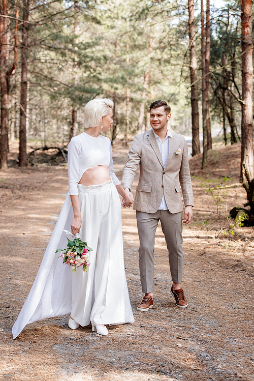 full length view of smiling just married couple holding hands and looking at each other in forest