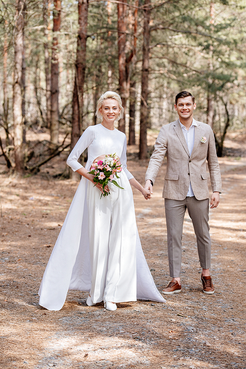 full length view of smiling just married couple holding hands in forest