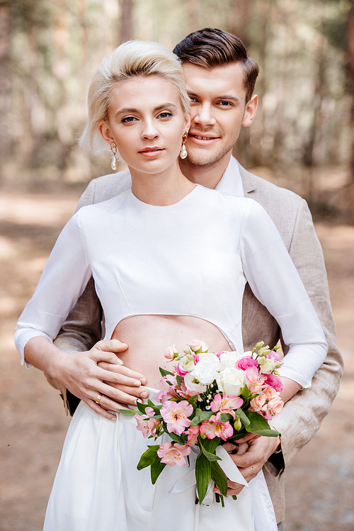 smiling married couple with wedding bouquet embracing in forest