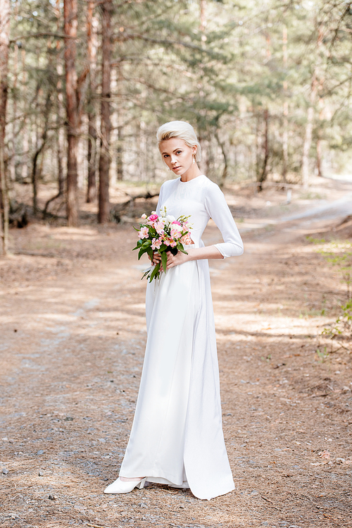 full length view of beautiful bride holding wedding bouquet in forest