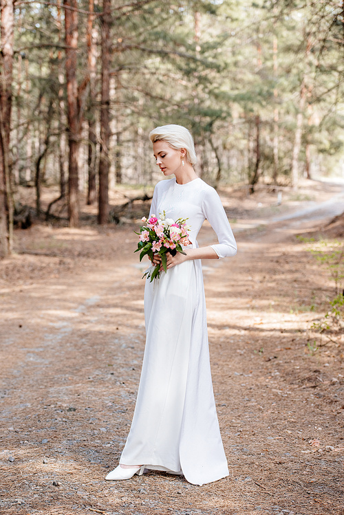 full length view of beautiful bride holding wedding bouquet in forest