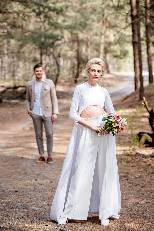 full length view of bride with bouquet of flowers and bridegroom in forest