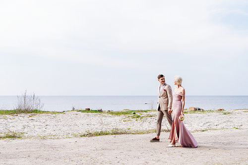 full length view of just married couple holding hands and walking on coast