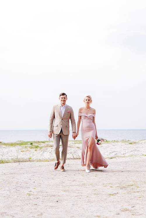 full length view of just married couple holding hands and walking on coast