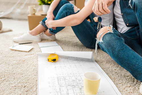 cropped view of couple sitting on carpet with blueprint, keys and measuring tape