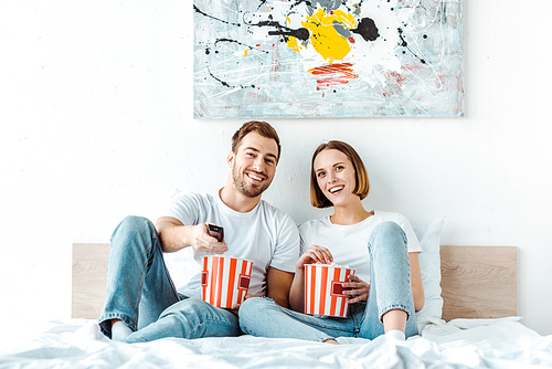 Joyful smiling couple eating popcorn in bed and watching tv