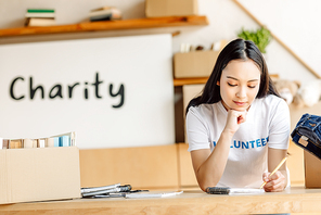 thoughtful asian volunteer writing in notebook while standing near carton boxes with clothes and books