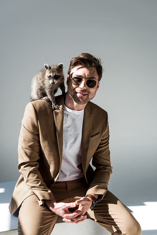 handsome man in sunglasses with cute raccoon on shoulder on grey