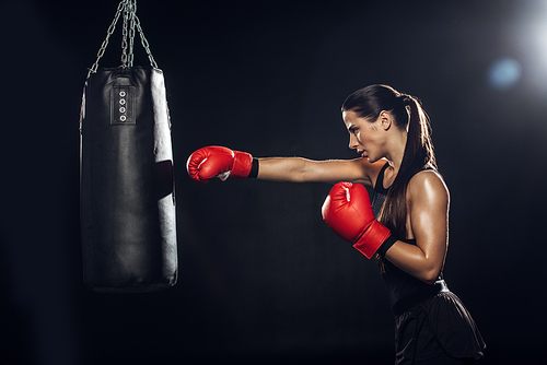 Side view of female boxer in red boxing gloves training with punching bag