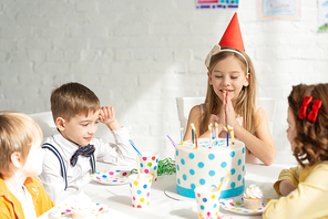 adorable kid doing please gesture and making wish while sitting at table with friends during birthday party celebration