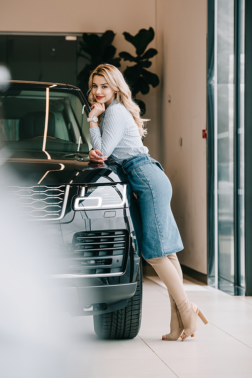 selective focus of happy blonde girl standing near black automobile in car showroom