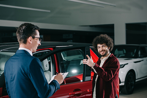 cheerful curly man looking at car dealer in glasses while standing near red car