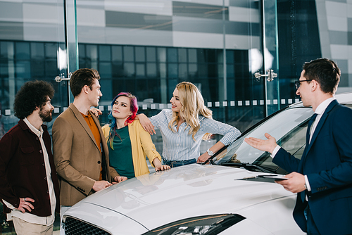 car dealer in glasses gesturing while looking at cheerful friends near white car