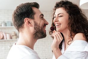 laughing curly woman feeding boyfriend with berry in kitchen