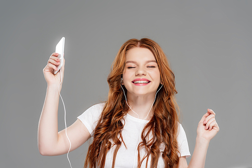 beautiful smiling redhead girl in earphones holding smartphone and listening music isolated on grey