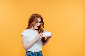 beautiful surprised redhead girl holding gift box isolated on yellow with copy space