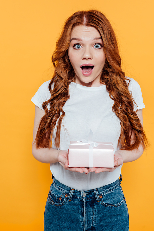 surprised redhead girl  and holding gift box isolated on yellow