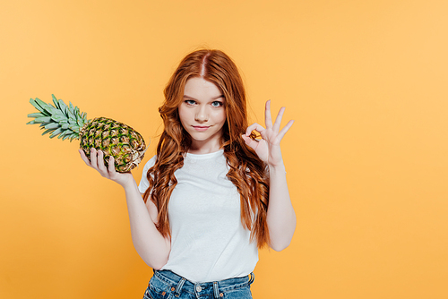 beautiful redhead girl  and showing ok sign while posing with pineapple isolated on yellow