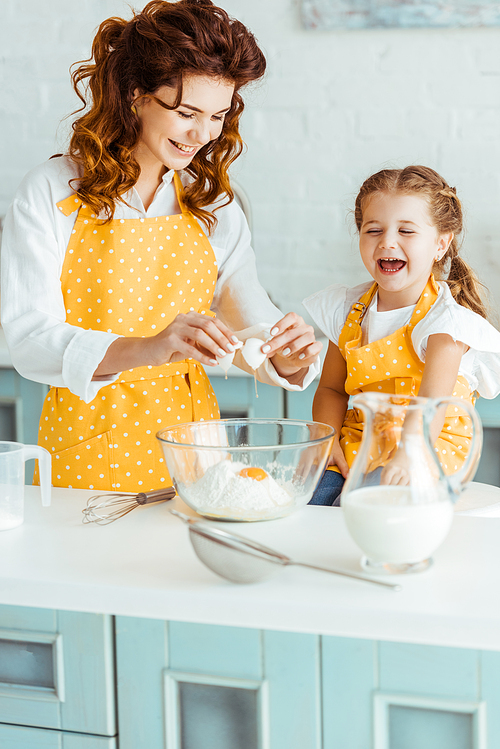 happy mother and daughter adding smashed egg to flour in bowl while cooking together in kitchen