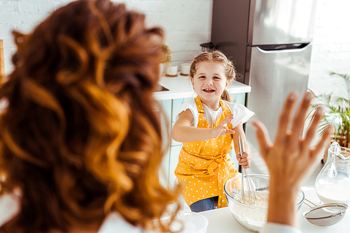 selective focus of excited kid in apron cooking dough in kitchen and looking at mother waving hand