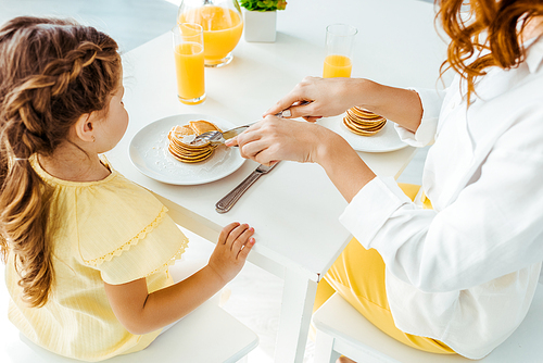 partial view of mother cutting pancakes with fork and knife near daughter
