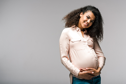 smiling pregnant african american woman touching belly while standing on grey