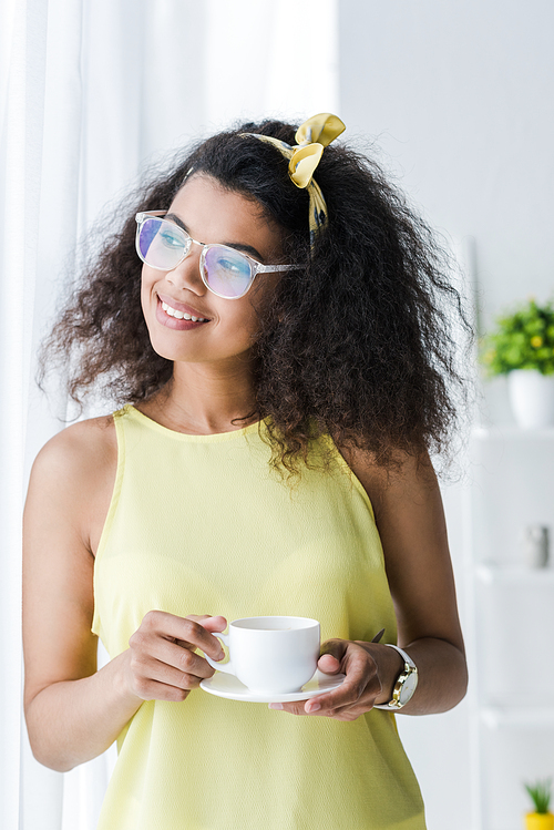 cheerful african american woman in glasses holding saucer and cup with drink
