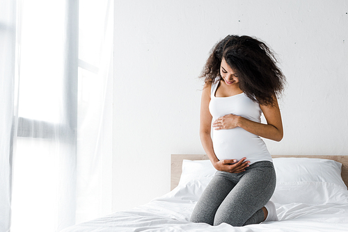 happy african american pregnant woman touching belly while sitting on bed