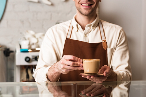 Cropped view of barista in brown apron holding cup of coffee with smile