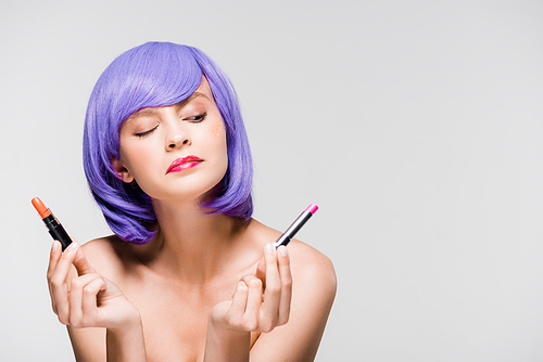 thoughtful young woman in purple wig looking at lipsticks isolated on grey