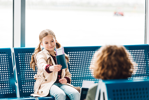 preteen kid sitting on blue seat, showing passport to boy in waiting hall in airport