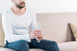 cropped view of bearded man sitting on couch and holding glass of water at home with copy space