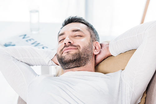 selective focus of handsome bearded man with eyes closed and Hands Behind Back resting on couch at home