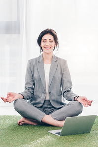 smiling businesswoman in suit meditating while sitting on grass mat in Lotus Pose near laptop