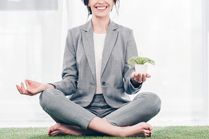 cropped view of smiling businesswoman in suit meditating on grass mat in Lotus Pose with flowerpot