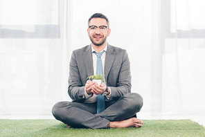 handsome smiling businessman in suit sitting on grass mat and holding plant in flowerpot