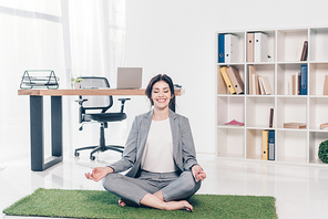 beautiful smiling businesswoman in suit sitting on grass mat in Lotus Pose and meditating in office