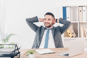 handsome smiling businessman in suit with Hands Behind Back sitting at desk in office