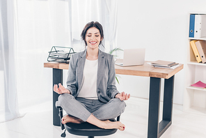 beautiful smiling businesswoman in suit sitting on chair and meditating in Lotus Pose in office