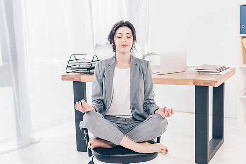 beautiful businesswoman in suit sitting on chair and meditating in Lotus Pose in office