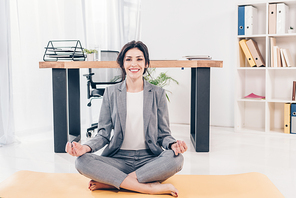 beautiful smiling businesswoman in suit sitting on fitness mat,  and meditating in office