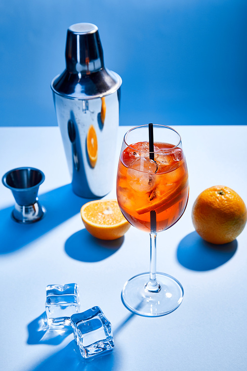 high angle view of cocktail Aperol Spritz, oranges, shaker, ice cubes and measuring cup on blue background