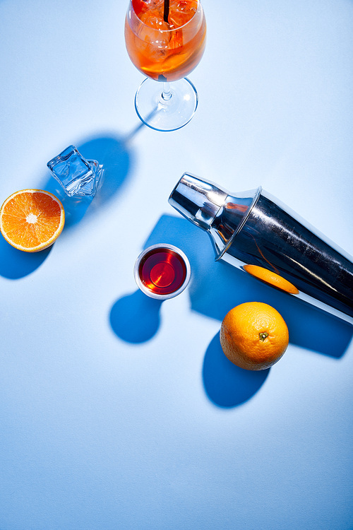 top view of Aperol Spritz, oranges, shaker, ice cubes and measuring cup on blue background
