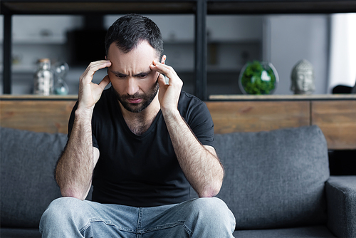 sad adult man holing hands near head while sitting on grey sofa at home