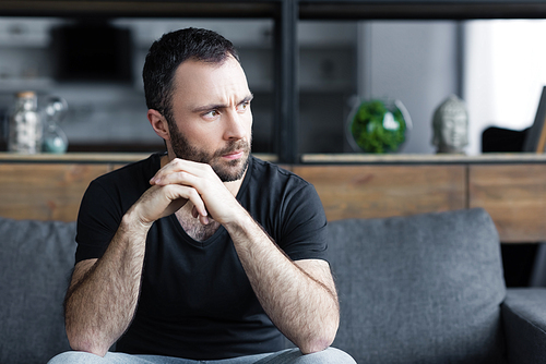 serious bearded man sitting on sofa with folded hands and looking away