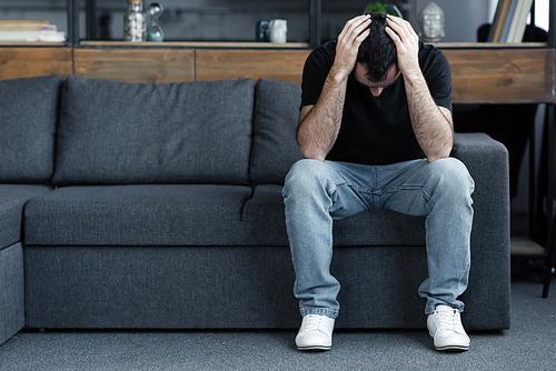 sad adult man in blue jeans sitting on grey sofa and holding hands on head